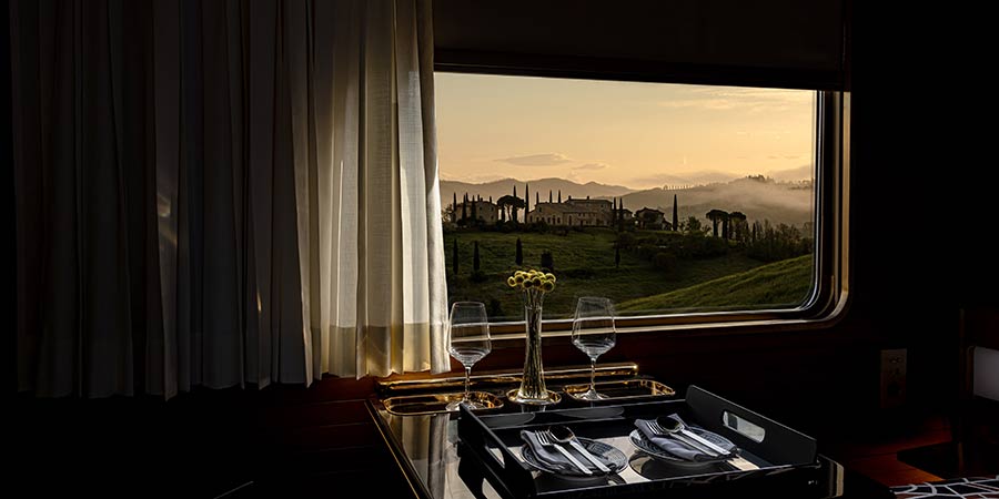 Looking out of La Dolce Vita Orient Express window with a curtain on the left with a view of traditional Tuscany buildings, rolling green hills and tall trees against a yellow sunset sky. In the carriage is a table with two dinner sets and wine glasses. 